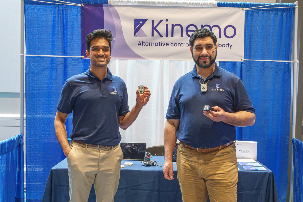 Two men in matching navy blue Kinemo polo shirts stand behind a blue branded booth, each holding a small rectangular device. A Kinemo banner hangs above them, with a table and additional booth materials in the background.