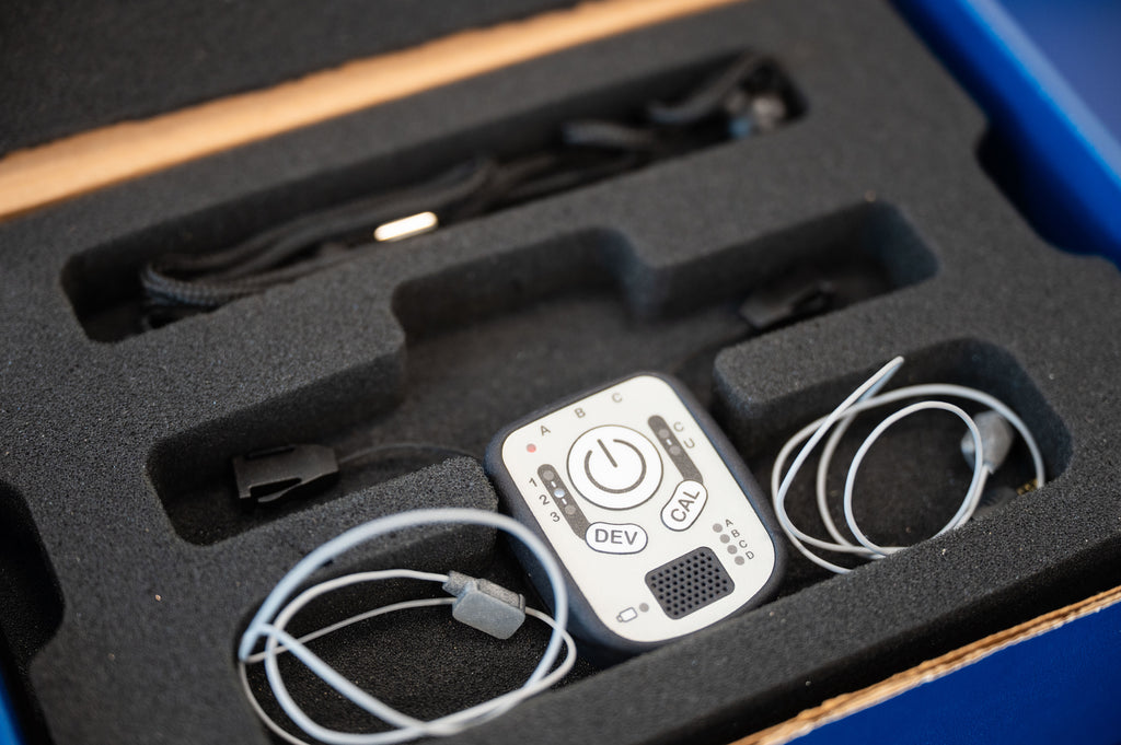 A close-up of a small white electronic device with printed buttons and labels, resting in a black foam tray alongside coiled cables inside an open box.