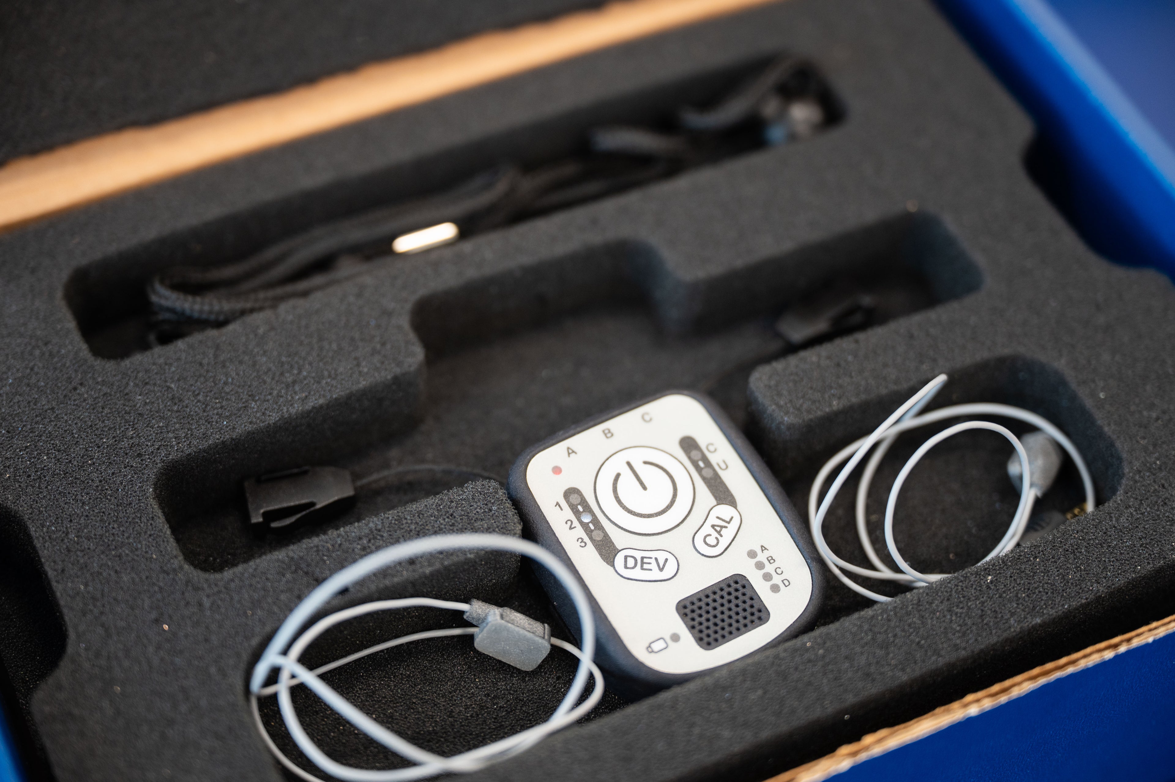 A close-up of a small white electronic device with printed buttons and labels, resting in a black foam tray alongside coiled cables inside an open box.