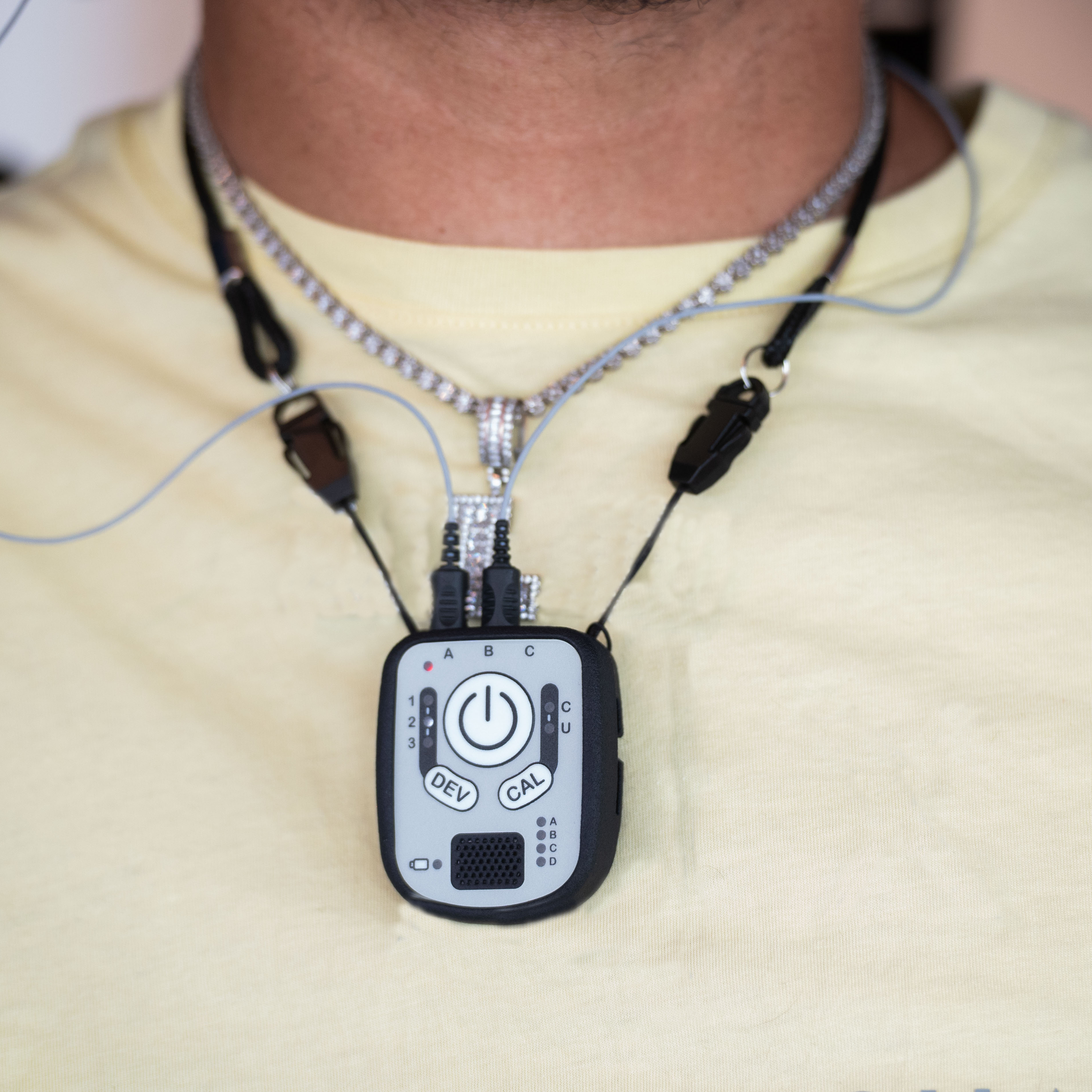 A close-up of a person wearing a silver chain necklace with a black and silver wireless control device hanging at the center of the chest.