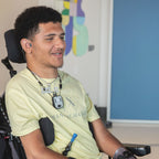 A man in a wheelchair in the center smiles while wearing a pale yellow shirt