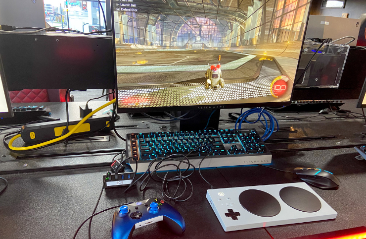 A retro gaming setup on a desk featuring a white Nintendo Classic Mini Console, a blue wireless controller, a mechanical keyboard with blue backlighting, and a monitor displaying a racing game.