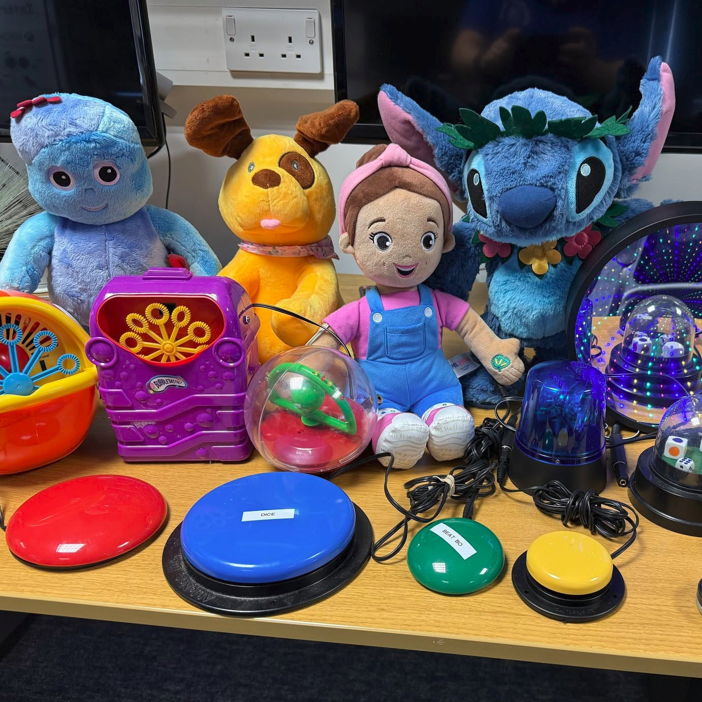 A collection of colorful special needs and sensory toys arranged on a wooden desk, including plush animals, buzzers, buttons, and a light box, with computer monitors visible in the background.