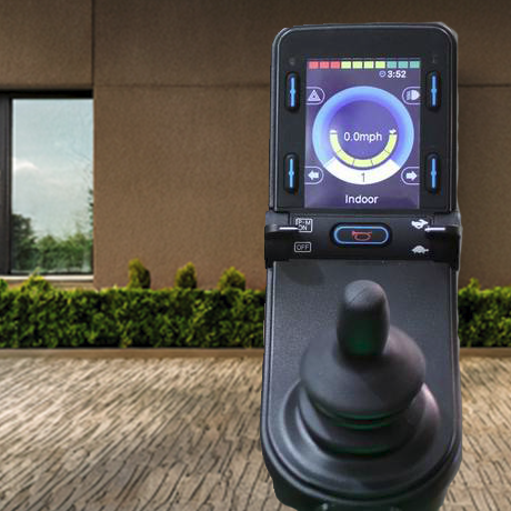 A black joystick control device with an attached display screen showing an indoor navigation interface, photographed outdoors in front of a modern building with a paved walkway and green hedges.