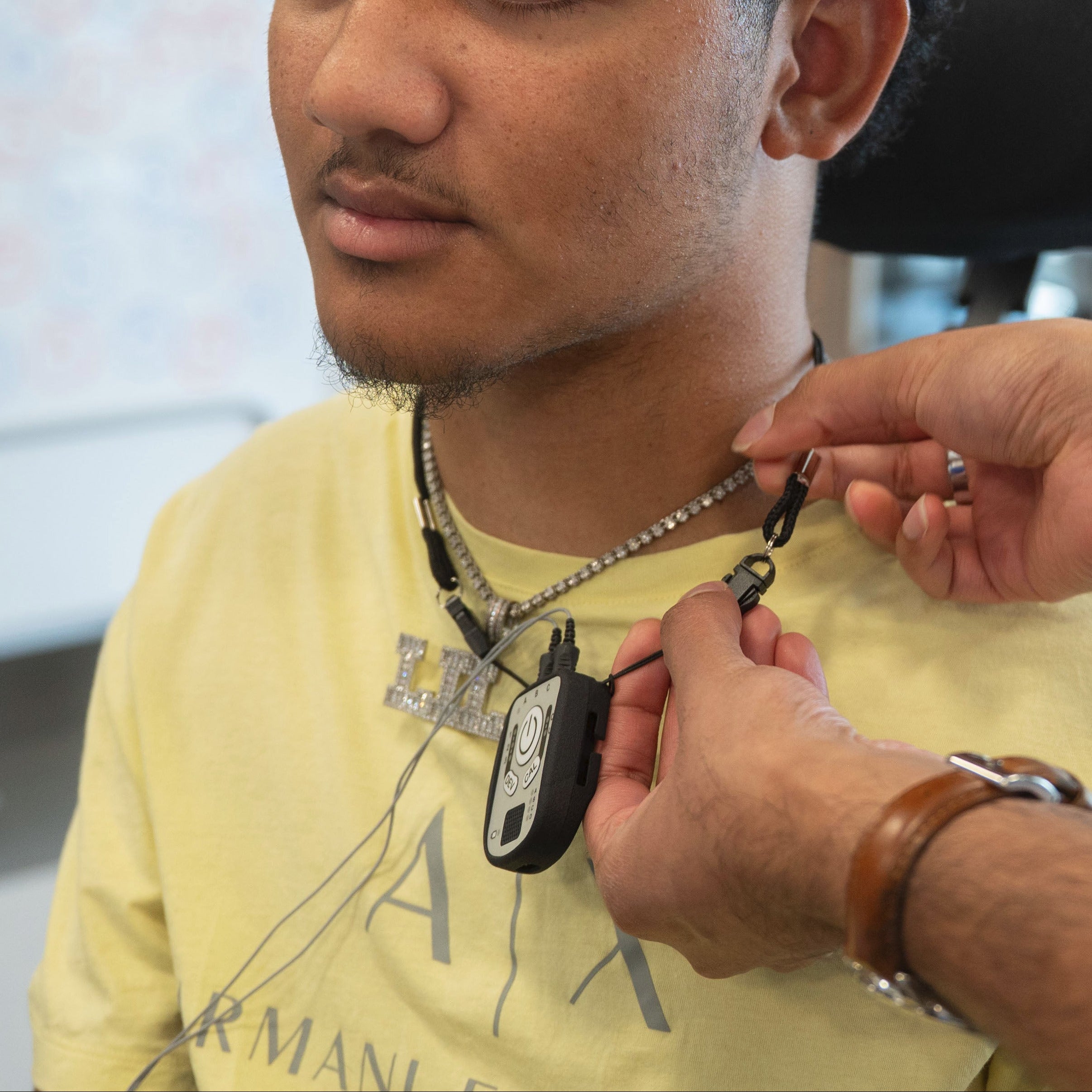 A man wearing a yellow t-shirt sits in a chair while another person adjusts a lanyard with a Kinemo Connect on his chest.