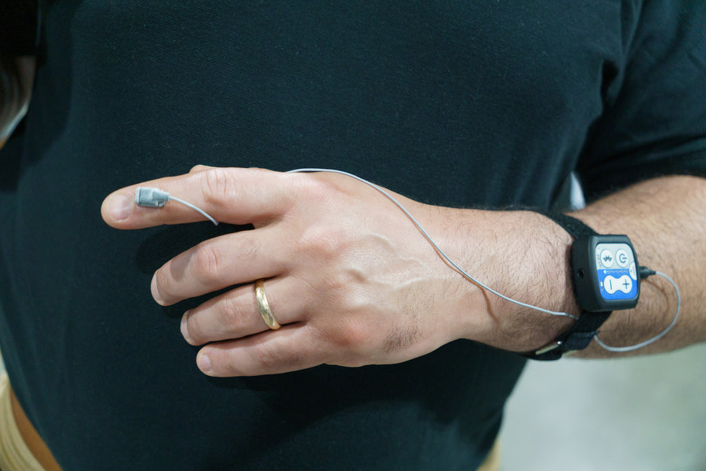 A close-up of a person's hand and wrist wearing a black electronic unit, connected to a thin wire leading to a small sensor on their index finger. The person is wearing a black t-shirt and a gold ring.