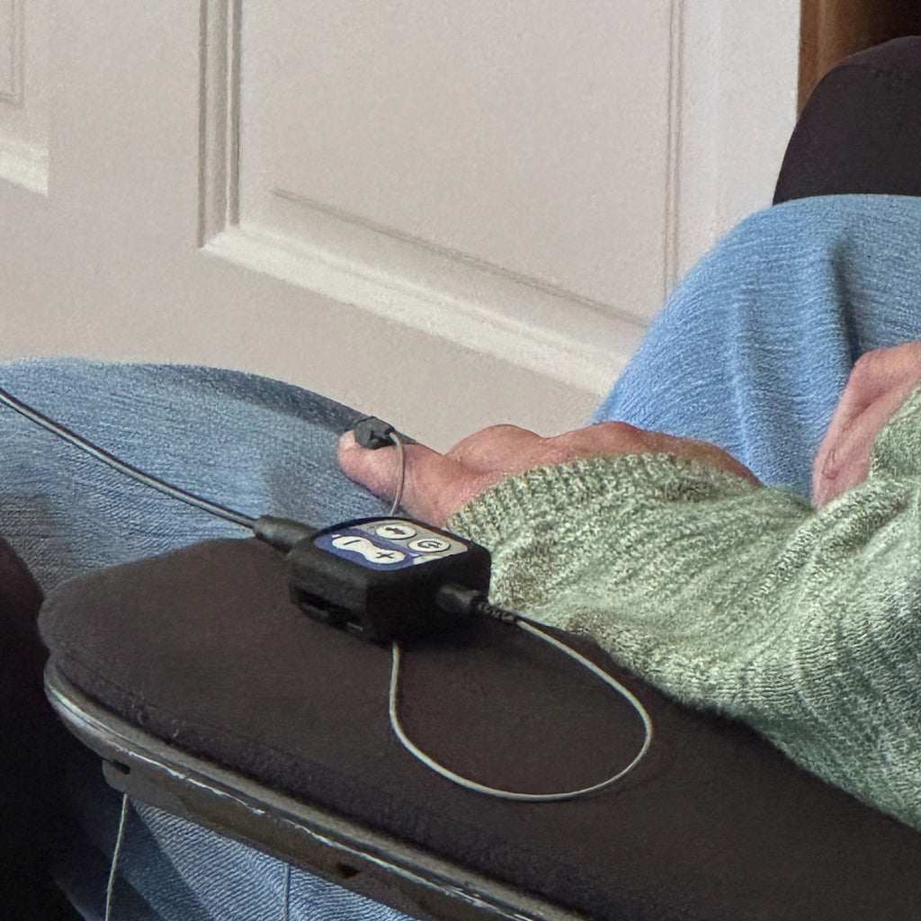 A close-up of a person's hand and arm resting on a dark chair cushion, wearing a small black electronic device connected by wires. The person is wearing a light green knit sweater and blue jeans, with a white paneled door in the background.