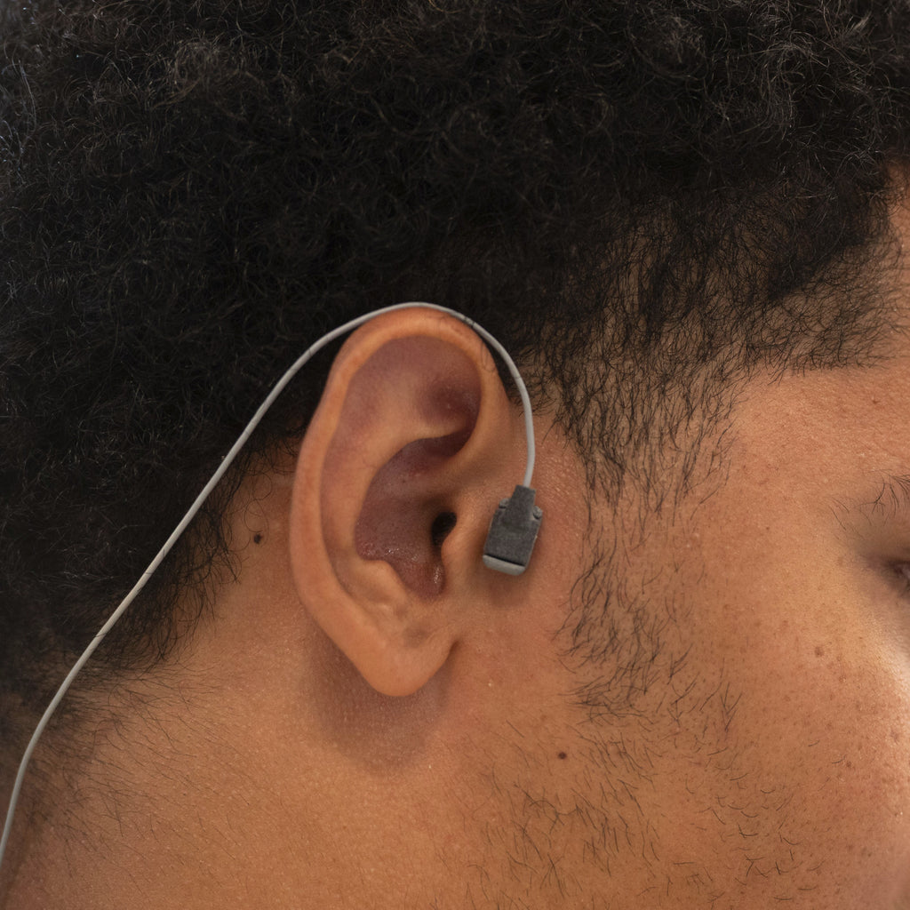 A close-up side profile of a man wearing a Kinemo tracer placed next to his ear with a thin wire extending from the tracer to his neck.
