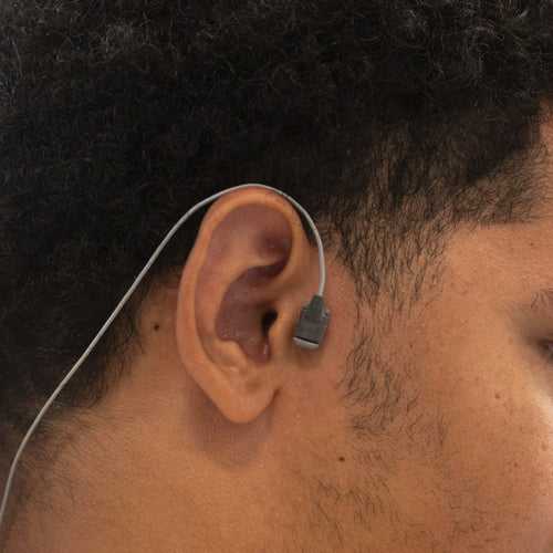 A close-up side profile of a man wearing a Kinemo tracer placed next to his ear with a thin wire extending from the tracer to his neck.