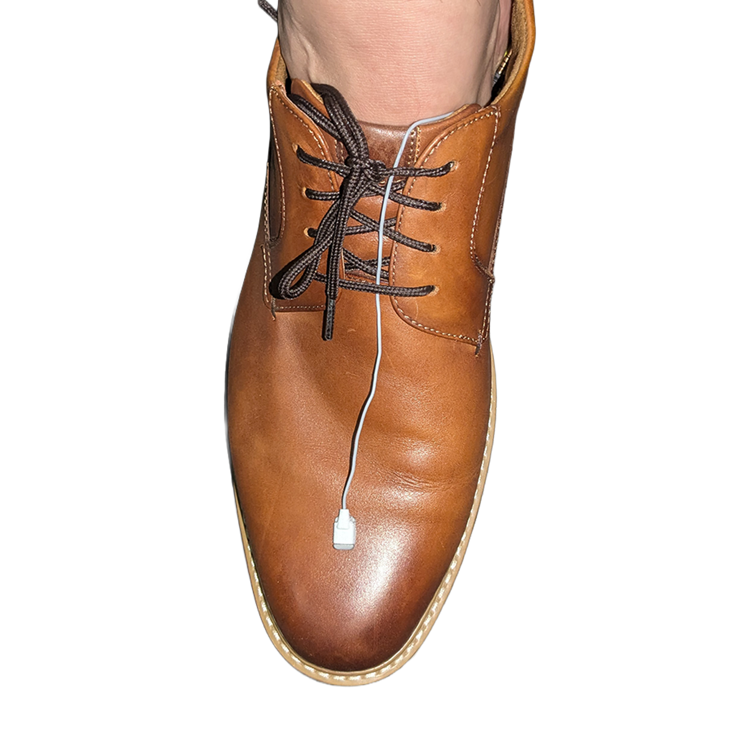 A close-up of a person's foot wearing a brown leather lace-up shoe, with a white cable threaded through the laces against a plain white background.
