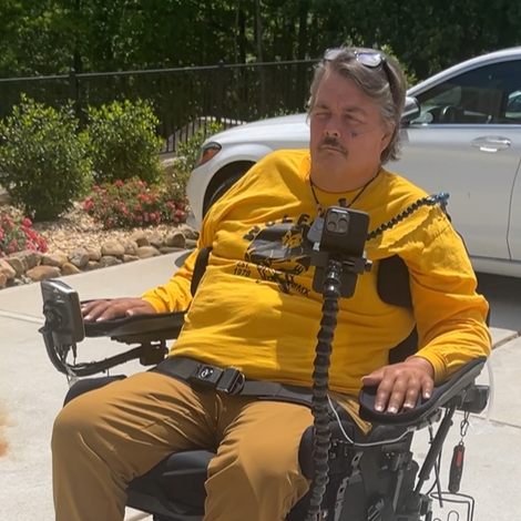 Person in a yellow shirt sitting in a wheelchair with a phone on a tripod outdoors.