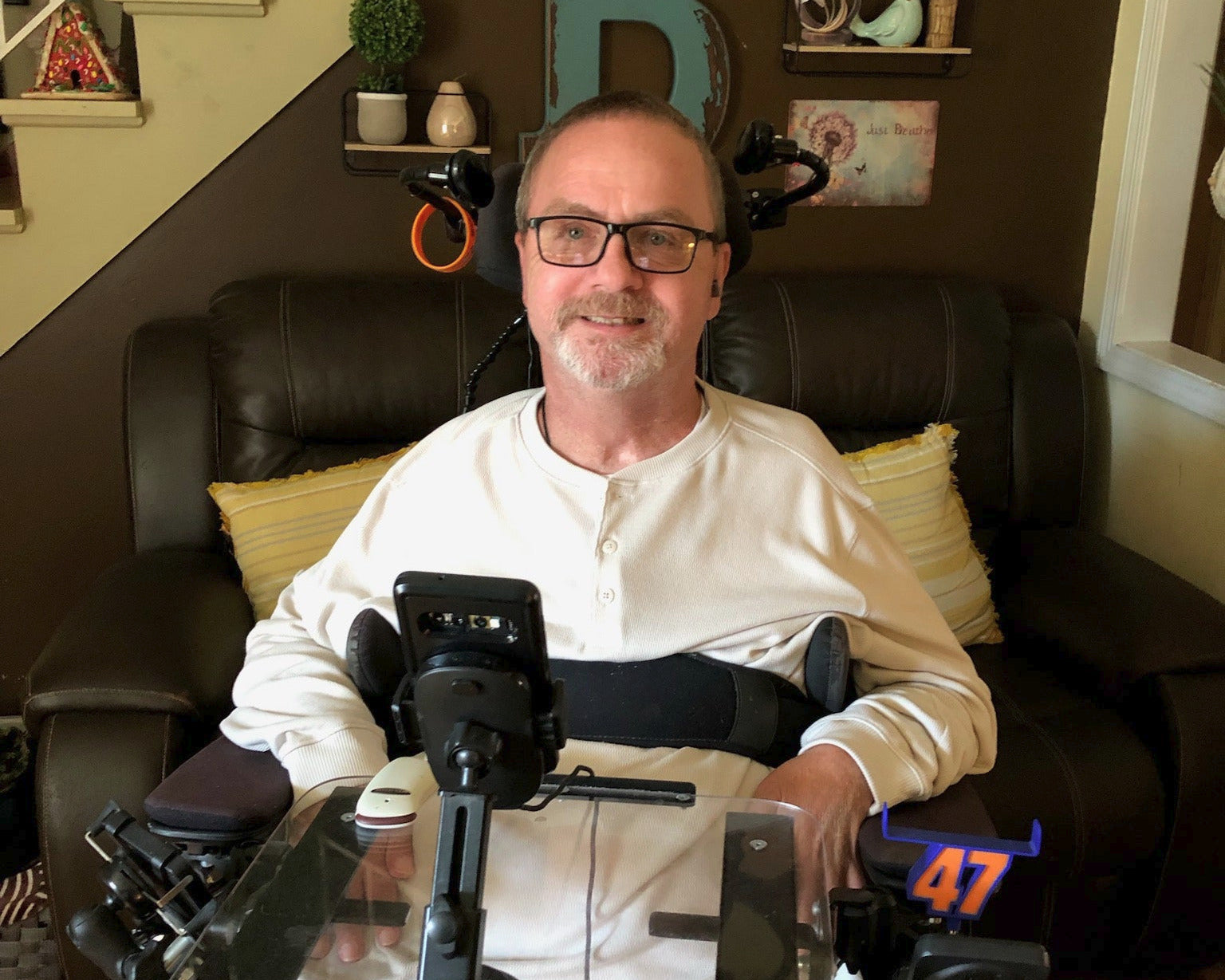 A man wearing glasses and a white henley shirt sits in a wheelchair with a clear tracking pad and camera setup in front of him, positioned in a living room with a dark leather couch and staircase in the background.